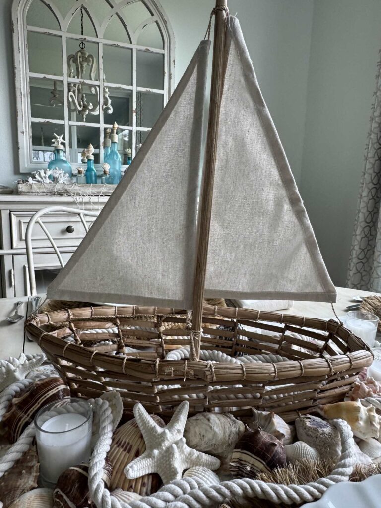 A woven decorative sailboat centerpiece with a white sail is surrounded by seashells, starfish, rope, and candles on a table, creating charming coastal decor. In the background, a mirror reflects coastal-themed accents.