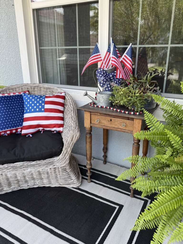 A wicker chair with a patriotic pillow sits beside a wooden table decorated with American flags, a potted plant, and red, white, and blue garland—perfect for showcasing your Patriotic Entertaining Ideas. A fern rests on a black and white rug in the foreground.