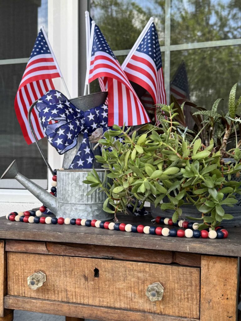 A rustic wooden table features a metal watering can with a blue star-patterned bow, small American flags, and a red, white, and blue beaded garland—perfect for showcasing your Patriotic Entertaining Ideas.