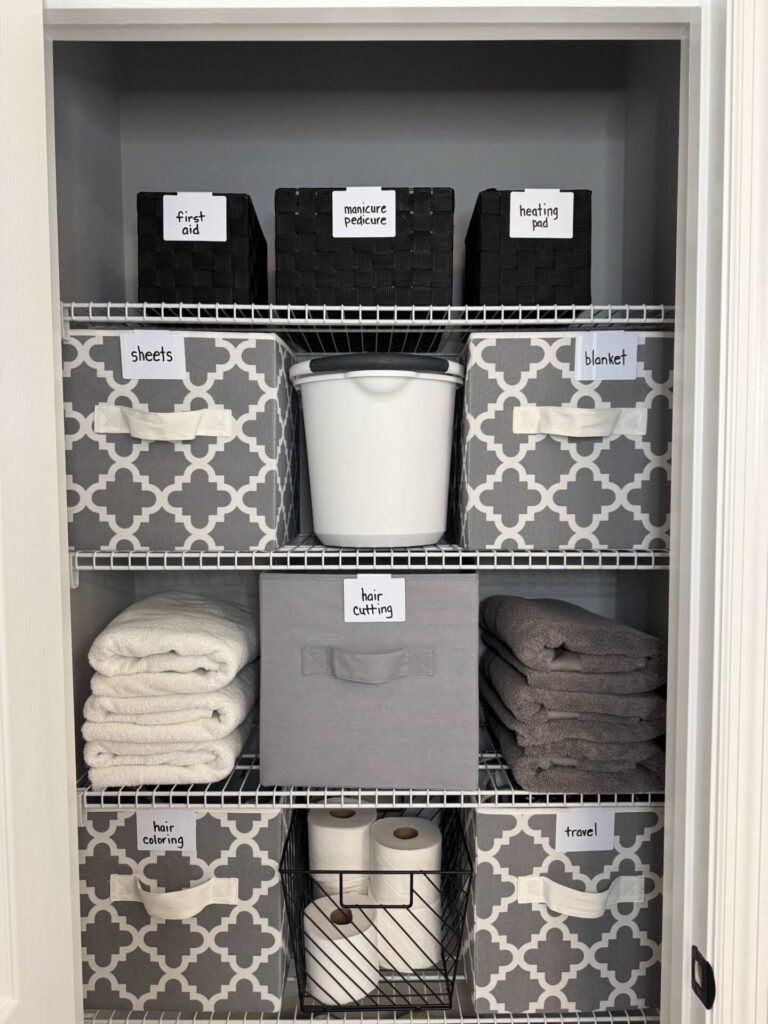 A neatly organized linen closet with labeled bins for first aid, manicure/pedicure, heating pad, sheets, blanket, hair cutting, hair coloring, travel items, towels, a white bucket—and a spot to organize greeting cards. Toilet paper and basket sit on the bottom shelf.