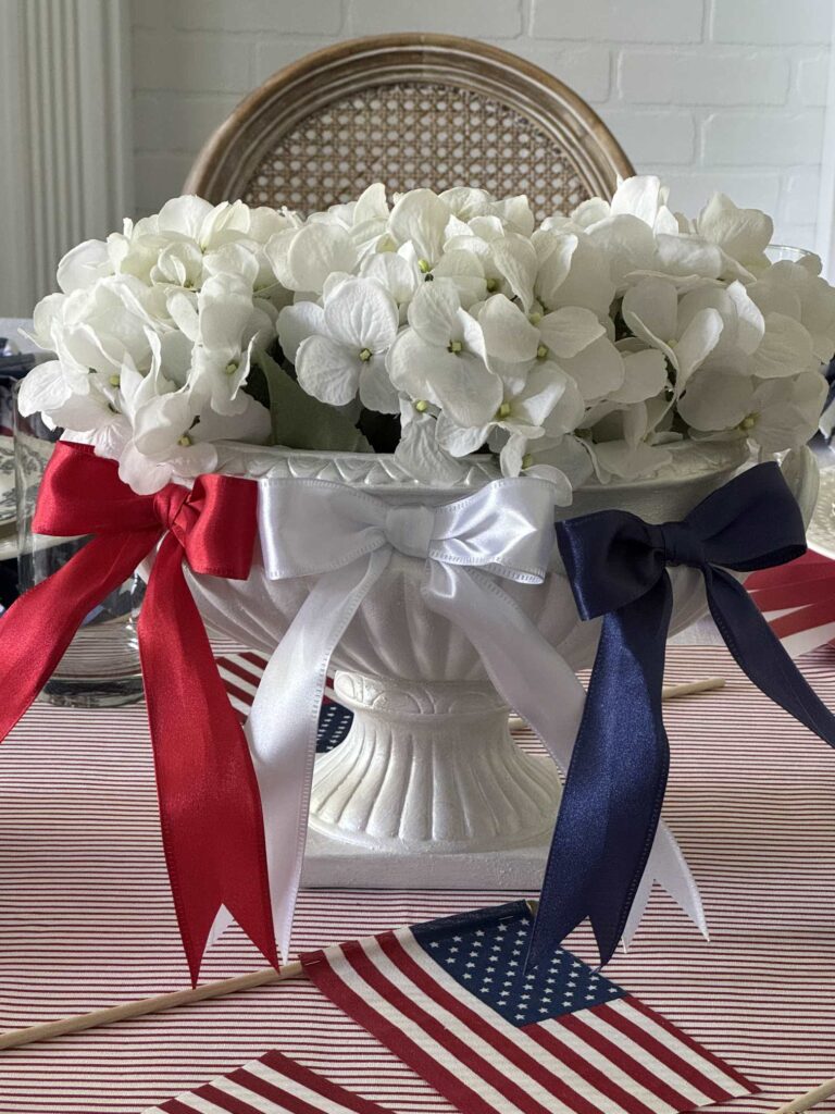 A white urn-style vase holds white hydrangeas adorned with red, white, and blue ribbons, sitting on a table with American flag decorations—perfect for showcasing your Patriotic Entertaining Ideas.