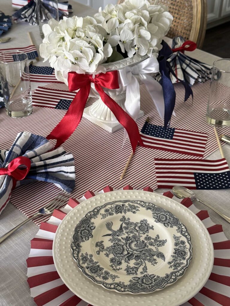 A dining table set for a patriotic celebration, featuring American flags, navy and white floral plates, a white floral centerpiece with red and blue ribbons—perfect for inspiring your next gathering with Patriotic Entertaining Ideas.