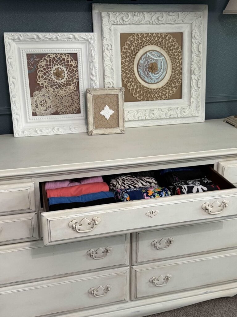 A white vintage dresser with ornate handles has a drawer open, neatly displaying folded clothes. On top, three framed doilies of different sizes and patterns are propped against a dark wall.