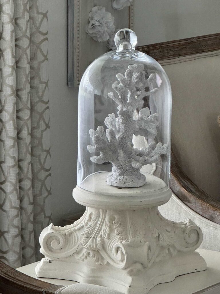 A white coral sculpture is displayed under a glass dome, set on an ornate ivory pedestal with scroll designs—an elegant accent for patriotic entertaining ideas—placed on a light-colored chair beside a patterned curtain.