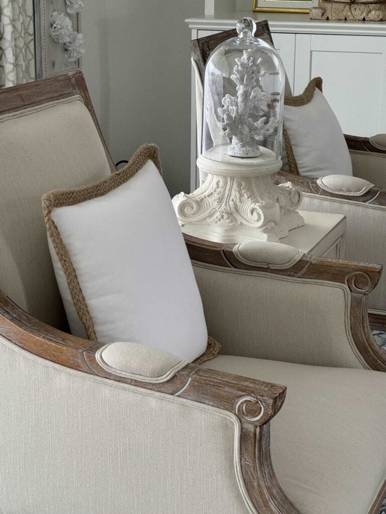 Two cream-colored armchairs with wooden trim sit beside a small white table. Part of the Seasonal Sentiments May 2026 Rewind collection, the chairs each feature a white cushion with tan rope edging and a glass cloche covers a white coral ornament.