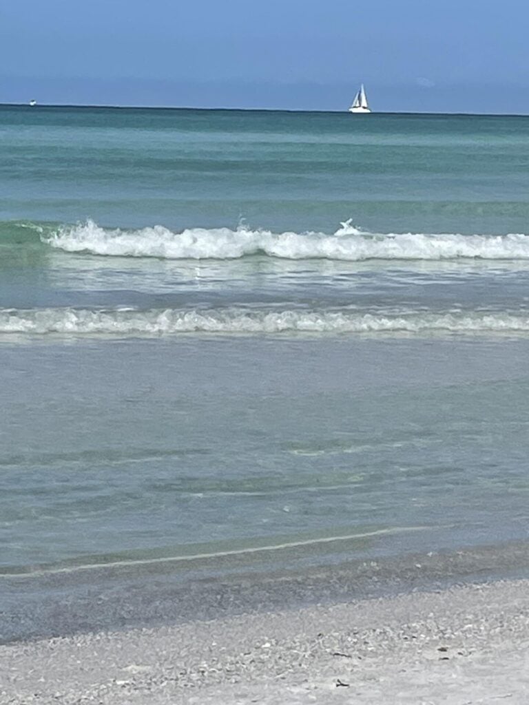 Light waves gently roll onto a sandy beach under a clear sky, with a white sailboat on the distant turquoise ocean—a scene perfect for reflecting on Seasonal Sentiments May 2026 Rewind.