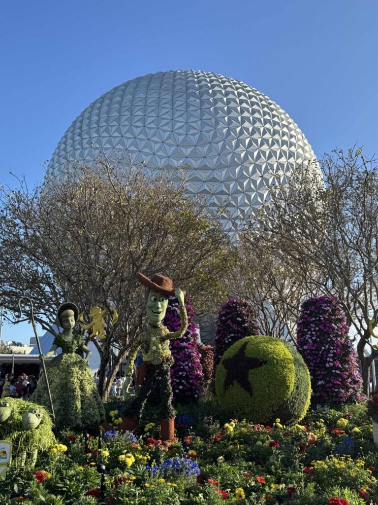 Topiary figures of Toy Story characters stand among colorful flowers in front of Epcot’s Spaceship Earth geodesic sphere, surrounded by trees under a clear blue sky.