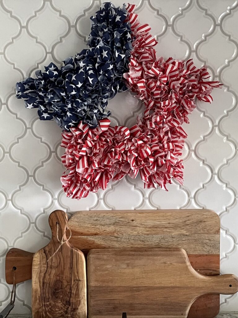A star-shaped wreath, perfect for Patriotic Entertaining Ideas, features blue and white stars on one half and red and white stripes on the other, hanging above a row of wooden cutting boards on a white tile wall.
