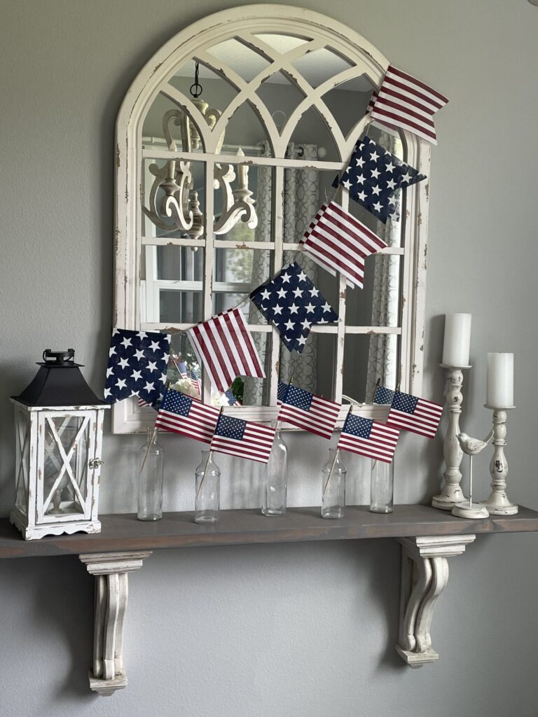 A decorative shelf with glass bottles holding small American flags sits below a white arched mirror. Perfect for Patriotic Entertaining Ideas, a garland of stars and stripes is draped across the mirror, with candlesticks and a lantern on the shelf.
