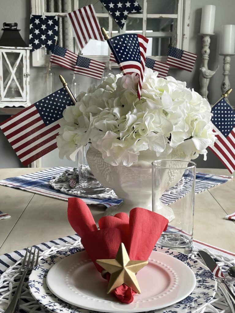 A table set for a patriotic celebration features white flowers in a vase with small American flags, plus a place setting with a red napkin and gold star—perfect inspiration for your Patriotic Entertaining Ideas.