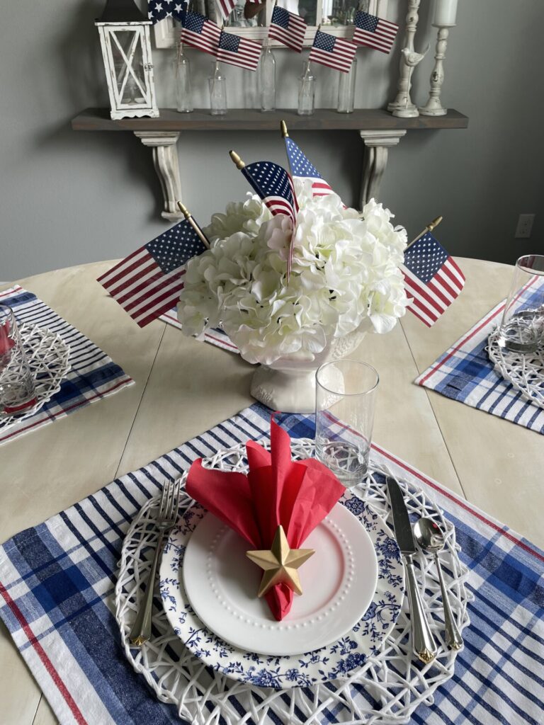 A patriotic table setting with red, white, and blue decor, featuring American flags, white flowers in a vase, a star on a red napkin, and plaid placemats—perfect for those seeking Patriotic Entertaining Ideas. Small flags are displayed in the background.