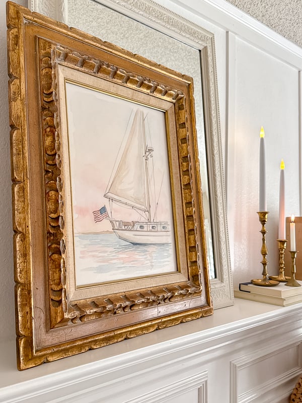 A gold-framed painting of a sailboat with an American flag is displayed on a white mantel beside brass candlesticks and books, reflecting the spirit of Seasonal Sentiments May 2026 Rewind in the large decorative mirror behind.