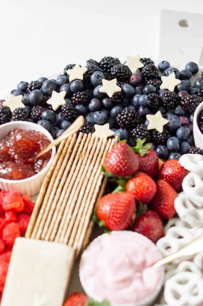 A platter featuring blueberries and blackberries topped with star-shaped cheese, strawberries, crackers, pretzels, fruit jam, and a bowl of pink dip.