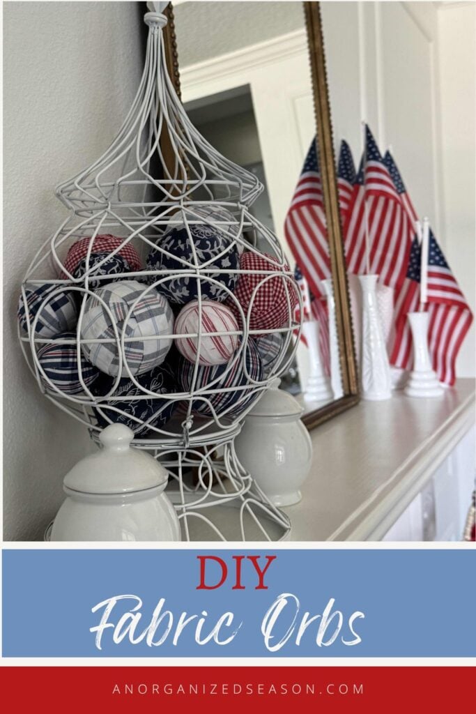 A white wire basket filled with DIY Fabric Orbs in red, white, and blue patterns sits on a mantel surrounded by white decor and several American flags in vases. Text below reads DIY Fabric Orbs - anorganizedseason.com.