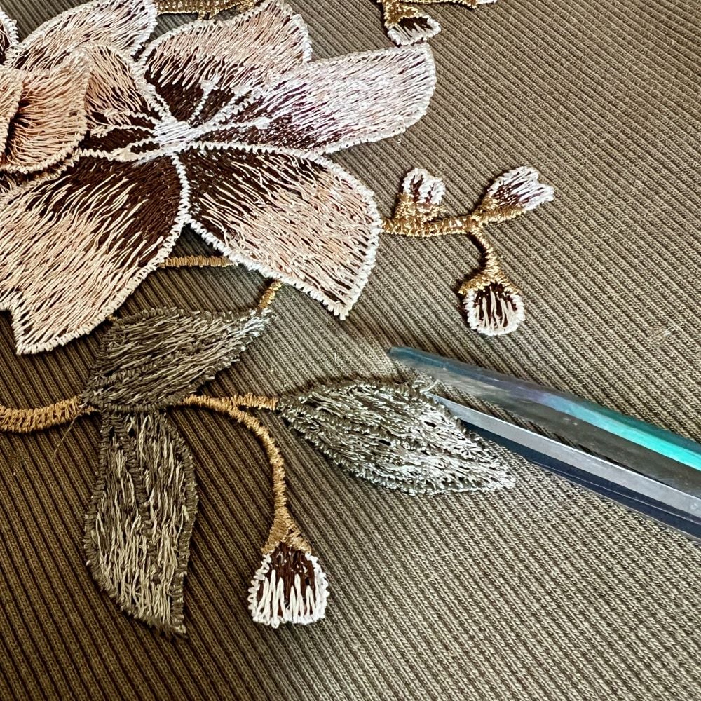 A pair of scissors is cutting around a large embroidered flower with leaves and buds on textured brown fabric.