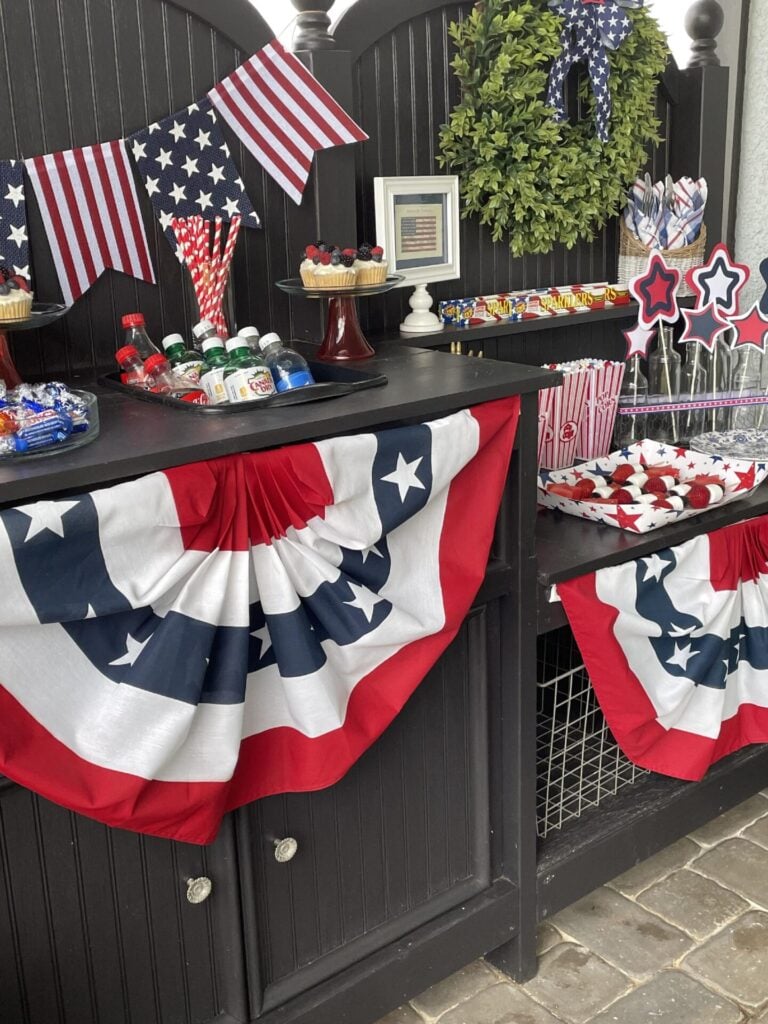 A festive display with red, white, and blue patriotic bunting, American flag decorations, drinks, cupcakes with berries, wreath, napkins, and star-shaped props set on a black cabinet.