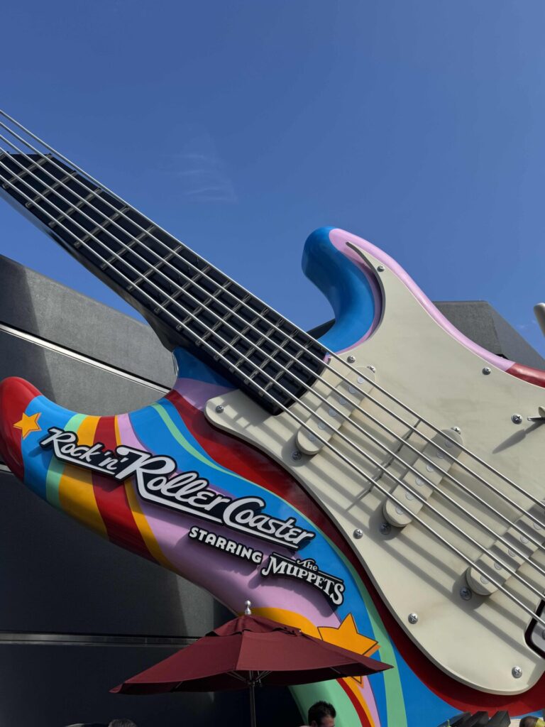 A giant, colorful electric guitar sculpture with rainbow stripes and the words Rock n Roller Coaster starring The Muppets is displayed outdoors against a blue sky and gray building. An umbrella is visible at the bottom.