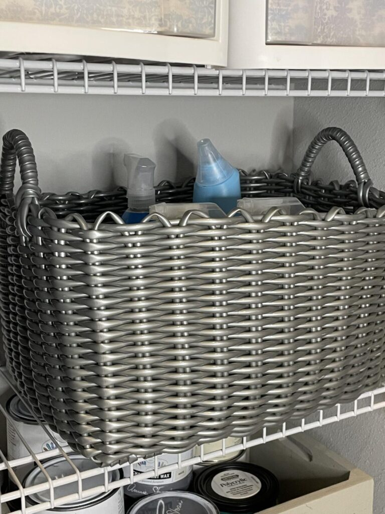 A gray woven basket with handles sits on a wire shelf, holding spray bottles and a blue-capped container. Below the shelf are cans of paint or similar products.