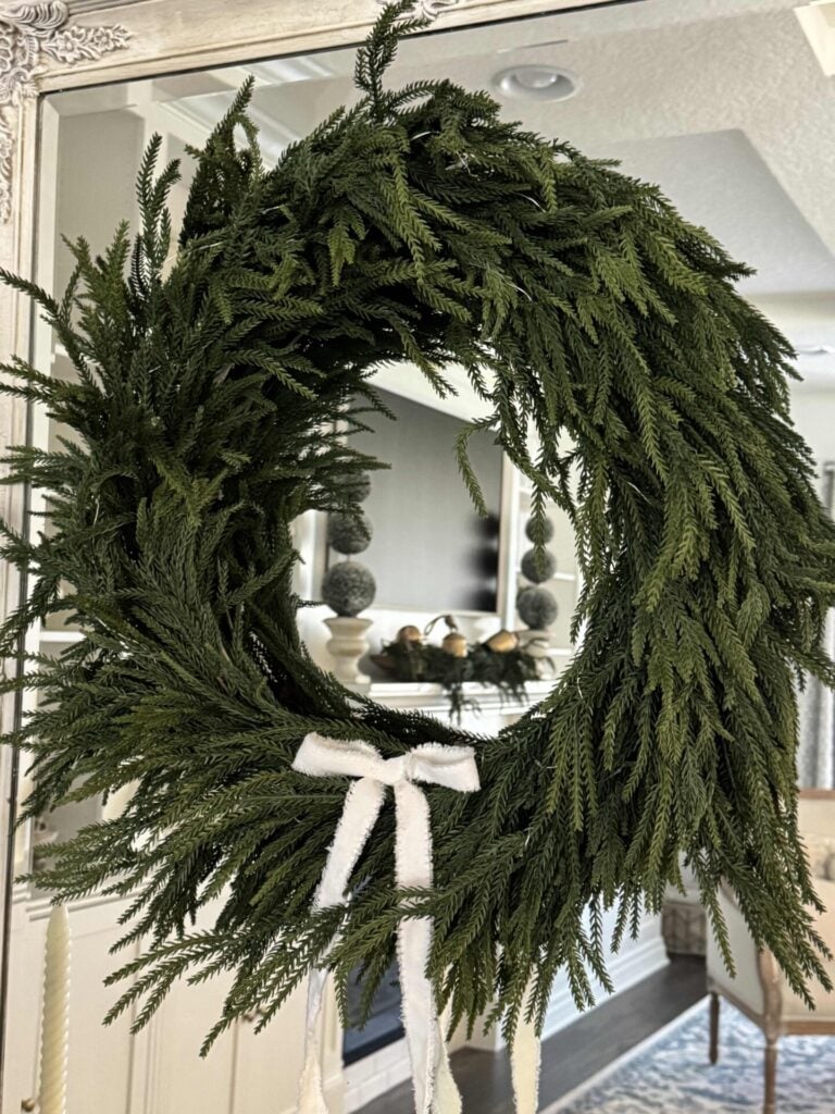 A DIY Frame Wreath of lush pine branches hangs on a mirror, adorned with a white fabric bow. The wreath’s reflection reveals a cozy, elegant living space decorated in peaceful neutral tones.