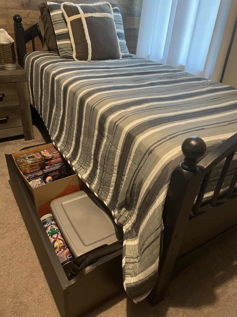 A neatly made bed with gray and white striped bedding sits next to a nightstand. An open drawer under the bed reveals storage containers, puzzles, and board games. Light streams through a window with sheer curtains.