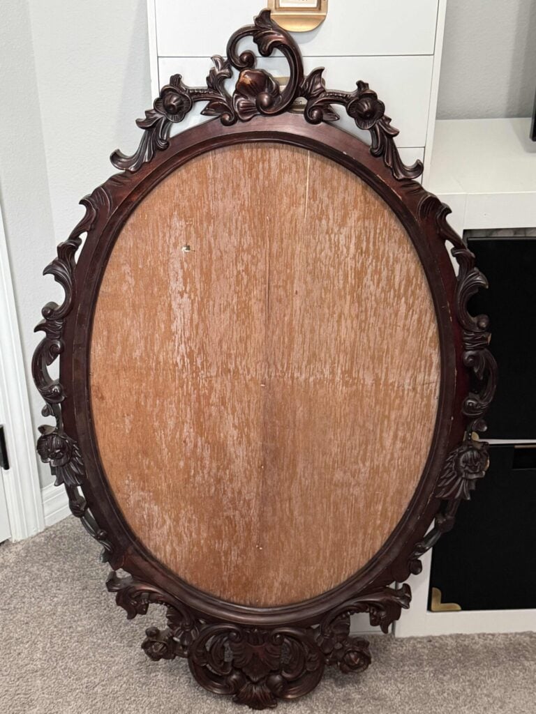 An ornate, empty oval wooden frame with intricate floral and leaf carvings rests against a wall on a carpeted floor. The frame has a reddish-brown finish and is missing its mirror or artwork. It's going to be the base for a DIY frame wreath.