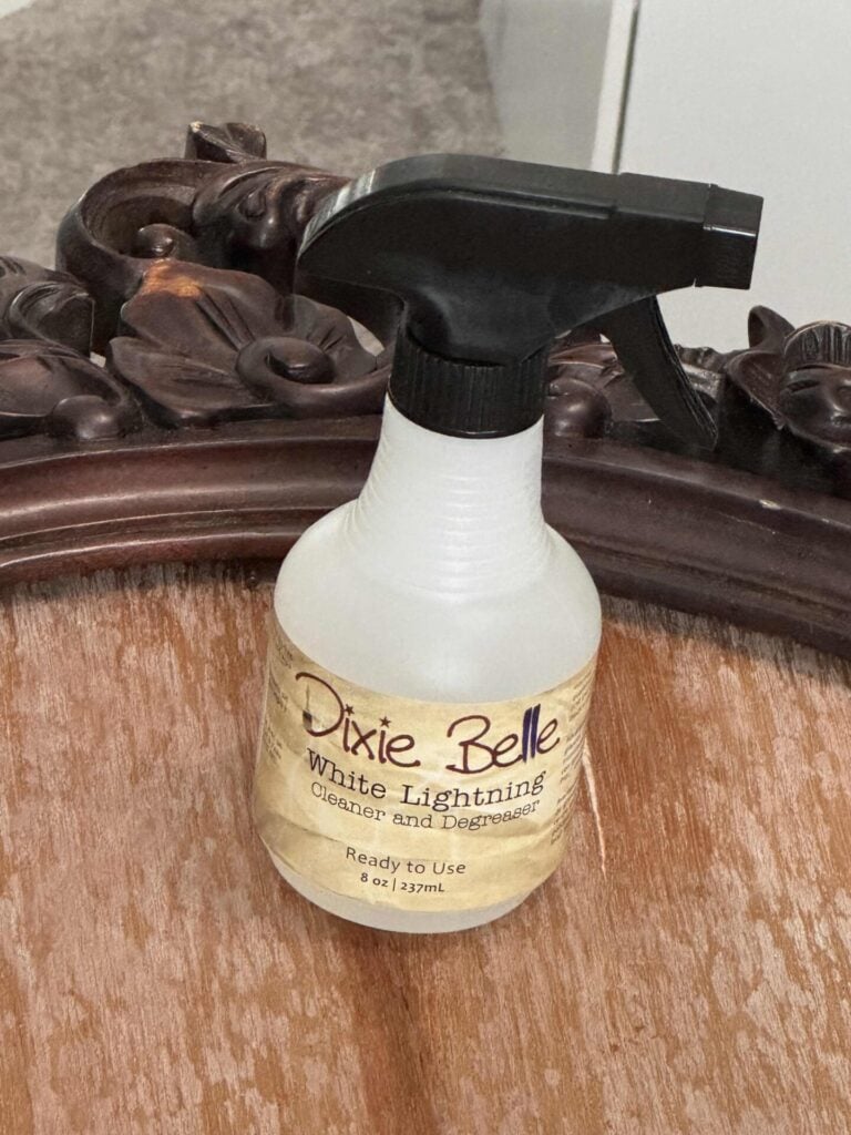 A spray bottle labeled Dixie Belle White Lightning Cleaner and Degreaser sits on a wooden surface next to an ornate, carved wooden mirror frame.