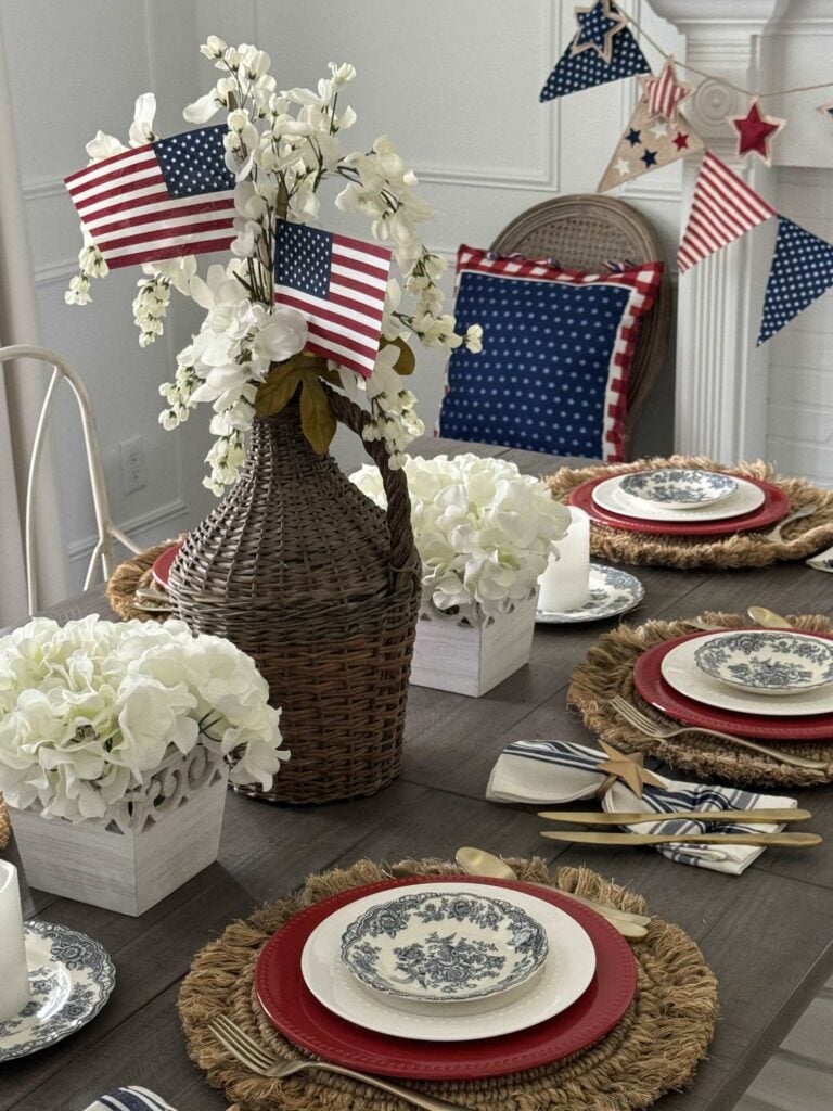 A dining table set for a patriotic celebration features American flags in a wicker vase with white flowers, blue and white patterned plates on red chargers, hydrangea centerpieces.