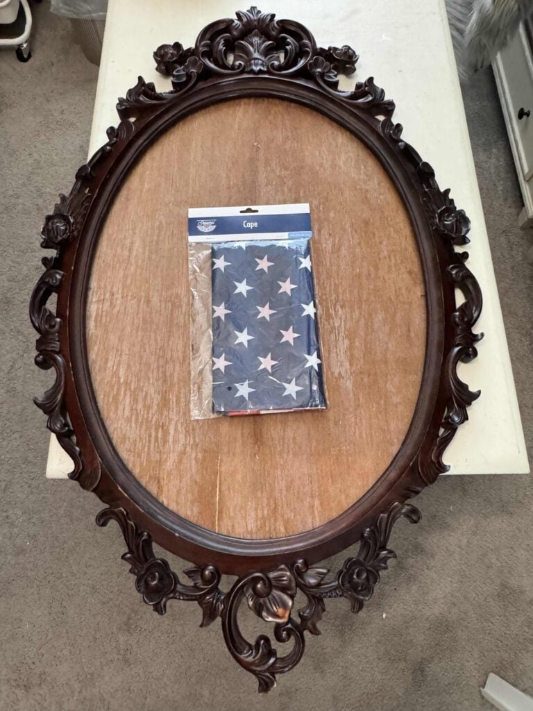 An empty, ornate oval wooden frame lies on a wooden surface with a folded American flag cape, still in its packaging, placed in the center. It's the base for a DIY frame wreath. 