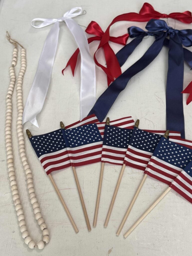 A set of small American flags on wooden sticks, a wooden bead necklace, and red, white, and blue ribbon bows arranged on a white surface.