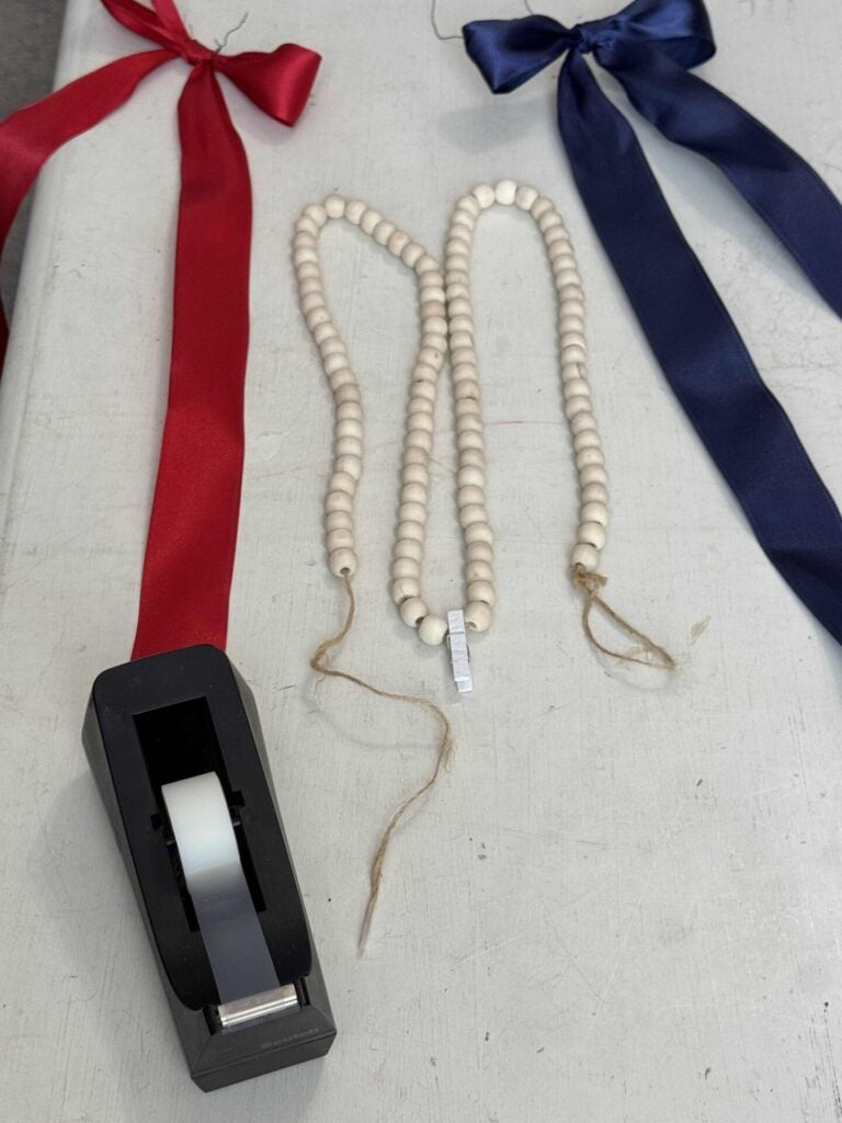 A roll of tape, a strand of wooden beads with twine ends, and wide red and navy blue satin ribbons are laid out on a white table.
