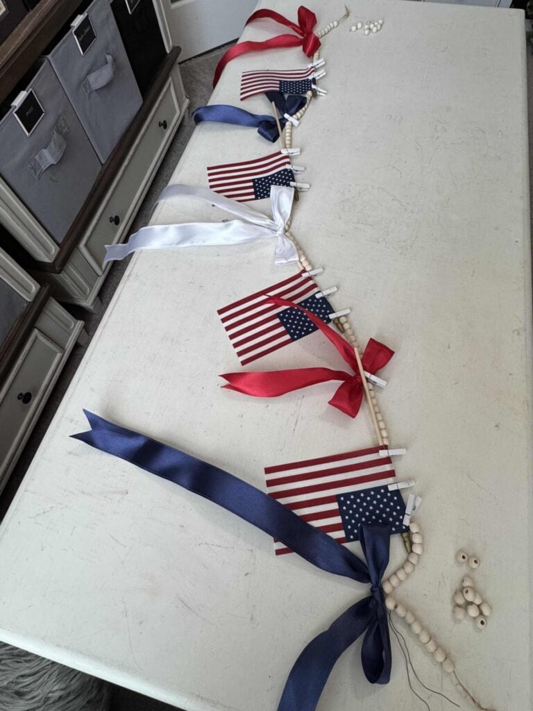 A tabletop displays a craft project with small American flags, red, white, and blue ribbons, and wooden beads strung together, likely for a patriotic decoration. Storage bins are visible in the background.