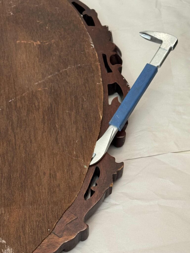 A metal pry bar with a blue handle is positioned between the edge of a wooden object with intricate cutouts and an off-white surface.