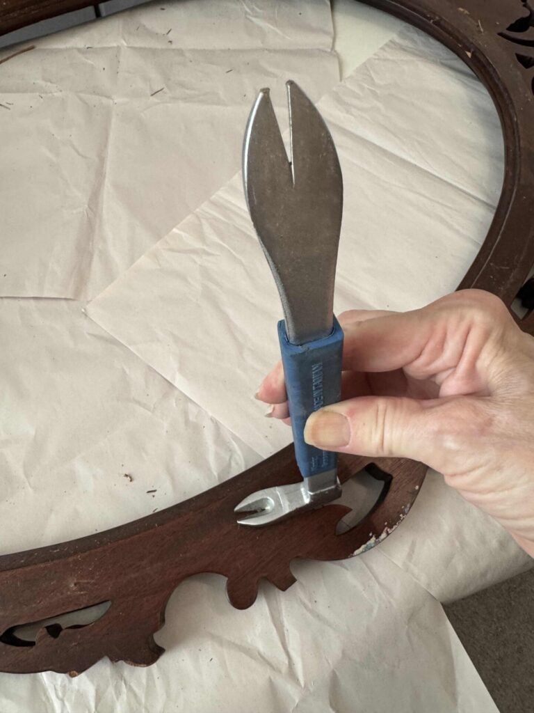 A hand holds a blue-handled tool with a wide, flat, forked metal end over an ornate wooden frame resting on a sheet of white paper.