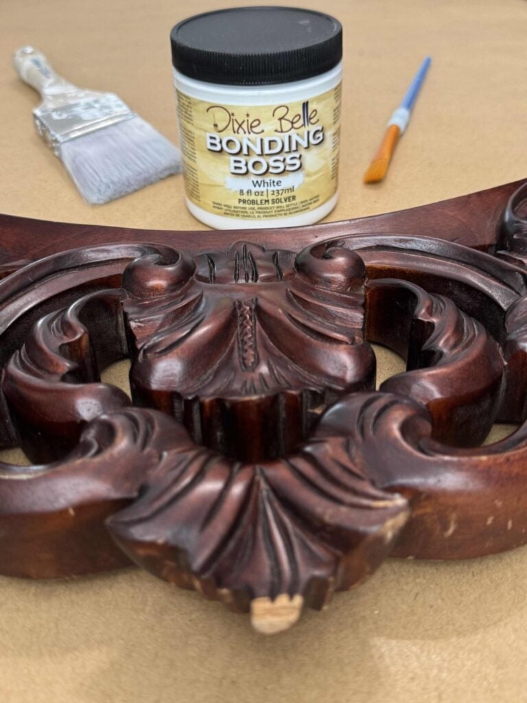 A carved wooden piece with a chipped edge sits in the foreground. Behind it are a jar of Dixie Belle Bonding Boss (White), a large paintbrush, and a small paintbrush on a brown surface.
