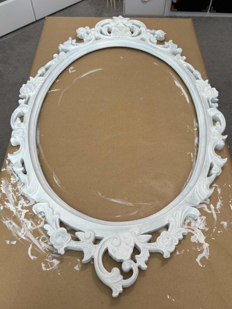 An ornate, white, oval picture frame with floral and scroll details rests on a brown work surface. White paint marks are visible on the surface near the bottom left of the frame.