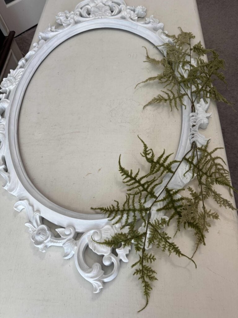 An ornate, empty white oval picture frame lies flat on a beige surface, decorated with sprigs of green fern leaves draped across its right side.