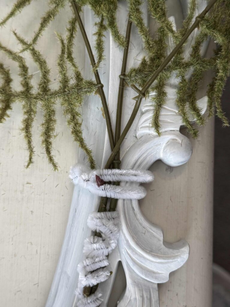 White pipe cleaners twist and secure green feathery stems to an ornate, white-painted decorative molding or frame, set against a light background.