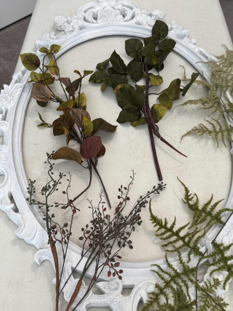 A decorative white oval frame lies flat on a surface, with various artificial greenery and branches with red berries arranged inside and around it.