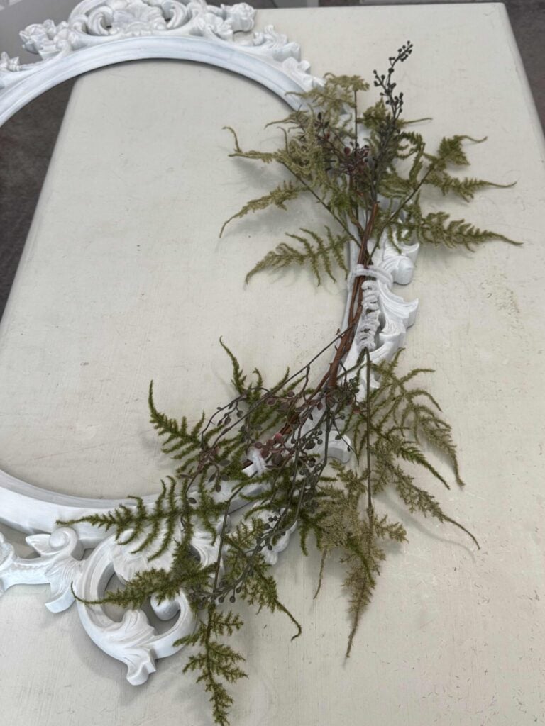 An ornate white oval picture frame lies flat on a light surface, decorated with green fern leaves and brown branches arranged along one edge, giving it a natural, rustic look.
