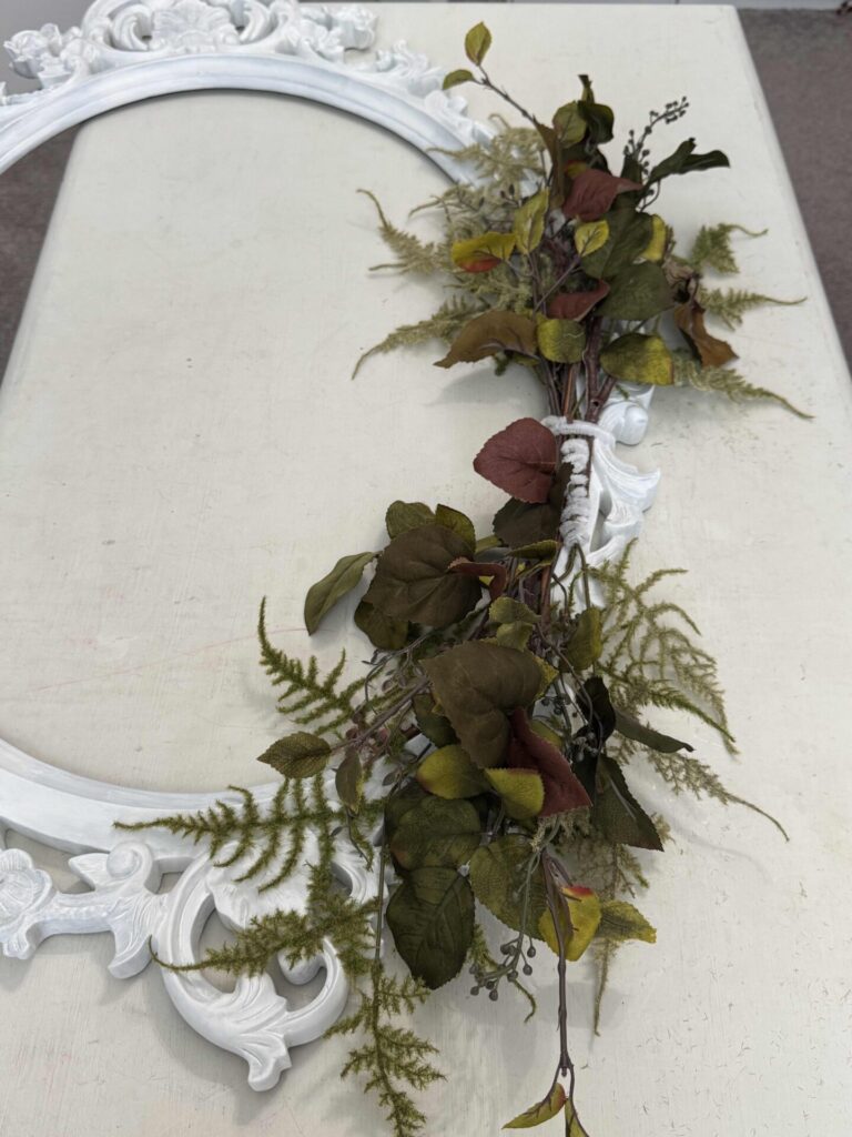 An ornate, white, empty picture frame lies flat on a surface, decorated with a garland of green ferns and brownish-green leaves draped diagonally across one side.