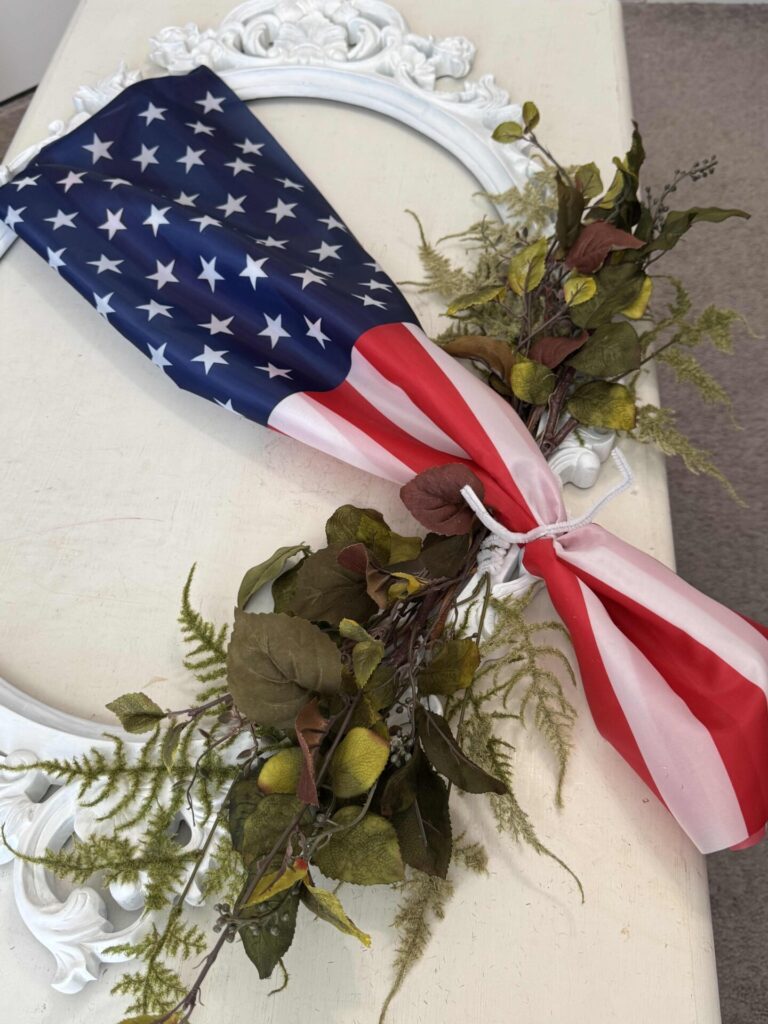 A partial American flag is tied to a white, ornate picture frame with artificial green leaves and branches arranged along the bottom edge of the frame, displayed on a light-colored surface.