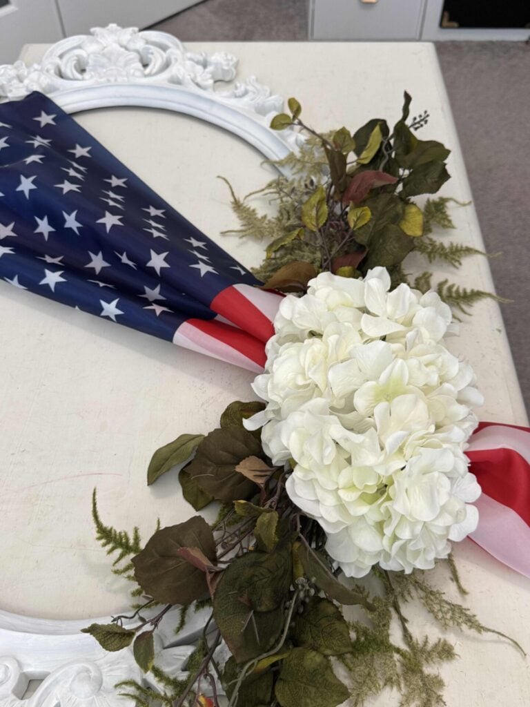 A decorative wreath featuring an American flag draped across a white ornate frame, accented with white hydrangea flowers, green leaves, and assorted greenery, resting on a light-colored surface.