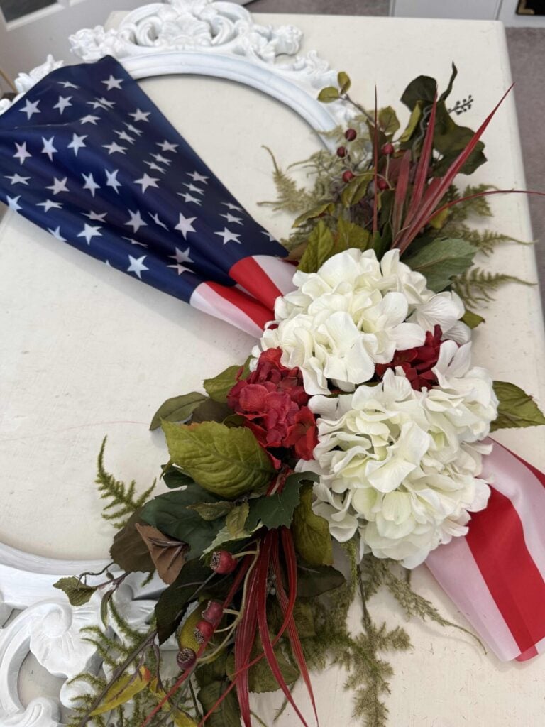 A wreath decorated with an American flag, white and red artificial flowers, green leaves, and sprigs of berries, displayed on a white surface with a white ornate frame in the background.