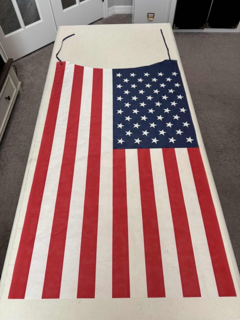 A white table displays an apron designed like the American flag, featuring red and white vertical stripes on one side and a blue field with white stars on the other.