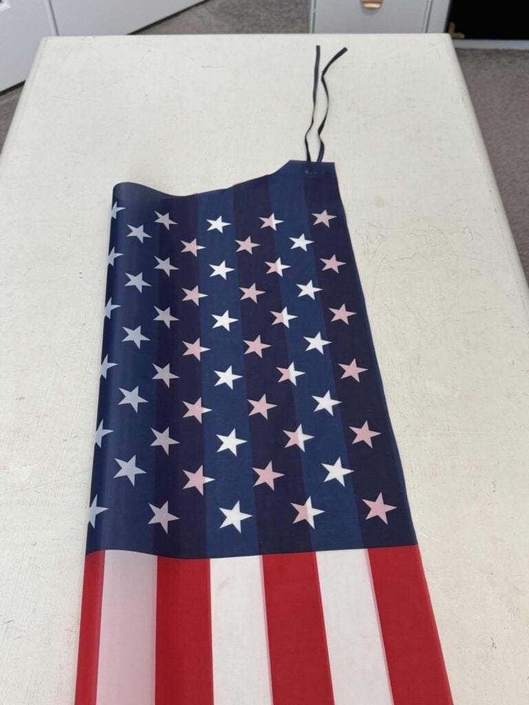 A fabric with a pattern of white stars on a blue background and red and white stripes lies flat on a white table. The fabric appears to be part of an American flag.