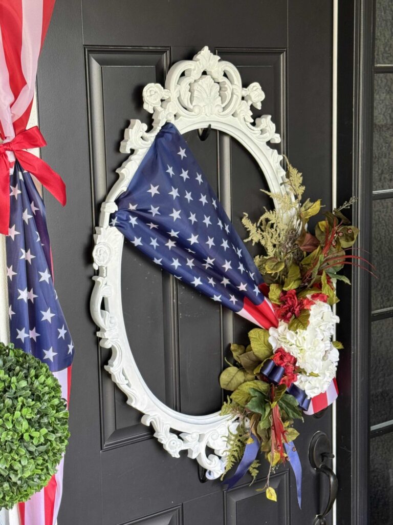 A decorative wreath on a black door features a white ornate frame, draped with an American flag and adorned with assorted greenery, red, and white flowers. Nearby, more patriotic fabric and a green topiary ball are visible.