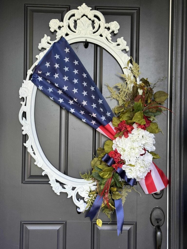 A decorative DIY Patriotic Ribbon Flag front door wreath features a folded American flag, white and red flowers, greenery, and blue ribbons arranged on an ornate white oval frame hanging on a dark door.