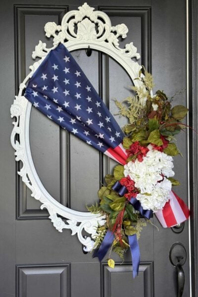 A decorative white oval frame wreath on a dark door, featuring a folded U.S. flag, white hydrangeas, red flowers, greenery, and blue and red ribbons arranged on one side for a patriotic display.