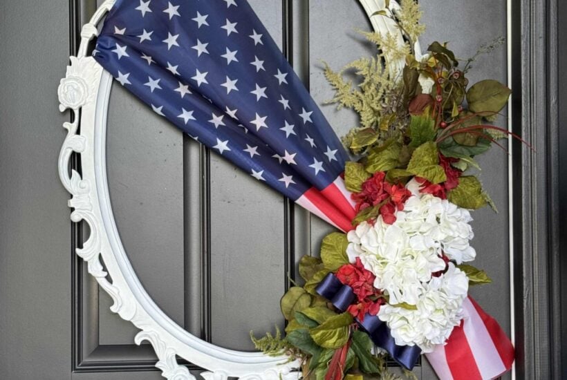 A decorative white oval frame wreath on a dark door, featuring a folded U.S. flag, white hydrangeas, red flowers, greenery, and blue and red ribbons arranged on one side for a patriotic display.