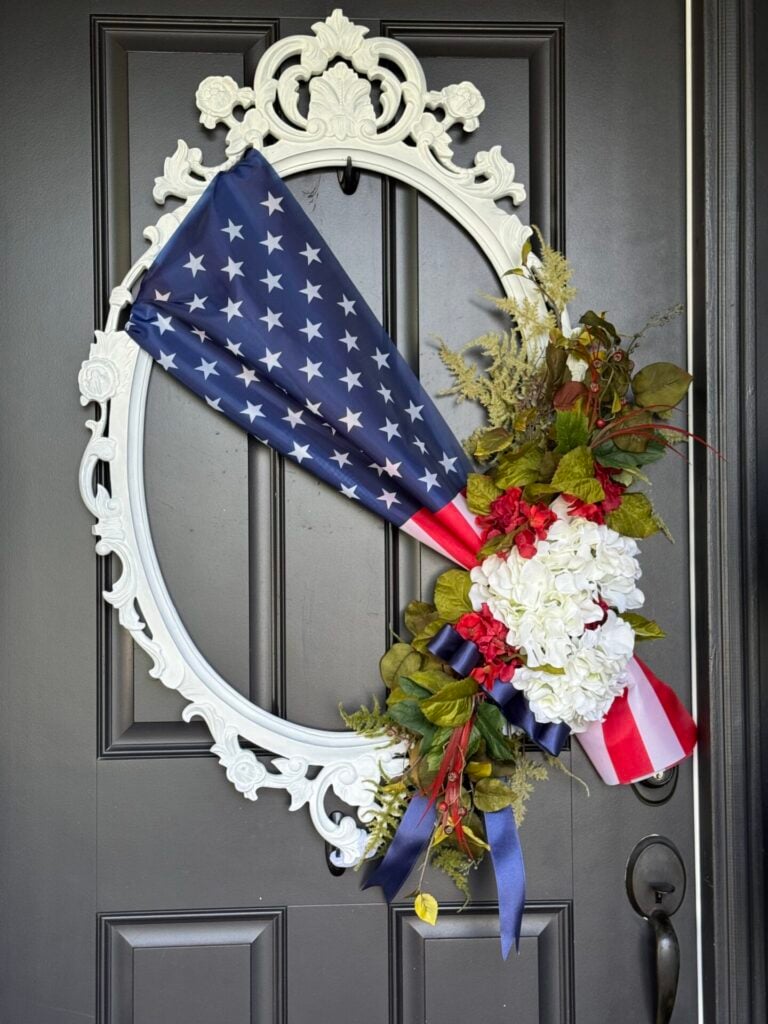 A decorative white oval frame hangs on a black door, displaying an arrangement with an American flag, white and red flowers, green foliage, blue ribbon, and a DIY patriotic garland.